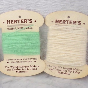 May include: Four spools of Herter's fly tying materials in various colors: dark brown, light green, off-white, and lavender. Each spool is mounted on a cardboard backing with the Herter's logo and text. The text reads "The World's Largest Makers and Dealers in Fly Tying Materials."
