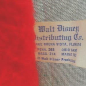 Puede incluir: Una etiqueta blanca con texto azul que dice "Walt Disney Distributing Co.". La etiqueta tambi&eacute;n incluye la direcci&oacute;n de la empresa: "Lake Buena Vista, Florida", y la informaci&oacute;n de contacto para Pensilvania, Ohio, Massachusetts y Maine.