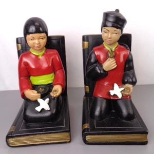 May include: A pair of bookends featuring two figures in traditional Asian clothing. The figures are kneeling and holding a white dove in their hands. The bookends are made of ceramic and have a black and gold finish.