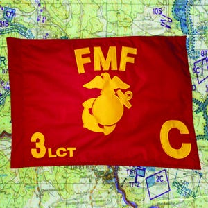 May include: A red flag with yellow lettering and a yellow emblem. The flag reads "FMF" above an emblem and "3 LCT C" below. The flag is set against a map background.