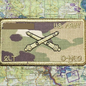 May include: A rectangular U.S. Army patch with a camouflage pattern. The patch features crossed cannons, the text "U.S. ARMY", and the text "2LT O-NEG". The patch is set against a map background.