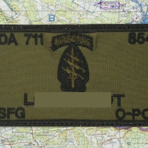 May include: Olive green rectangular patch with black embroidered text and a black emblem. The patch reads "ODA 711 AIRBORNE 8541" and "7SFG L T O-POS". The emblem is a black arrowhead with three lightning bolts.