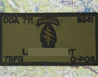May include: Olive green rectangular patch with black embroidered text and a black emblem. The patch reads "ODA 711 AIRBORNE 8541" and "7SFG L T O-POS". The emblem is a black arrowhead with three lightning bolts.