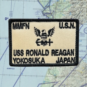 May include: A rectangular patch with black borders and beige background. The patch features the text "MMFN", "U.S.N.", "USS RONALD REAGAN YOKOSUKA JAPAN", and an emblem of an eagle above an anchor. The patch is set against a map background.