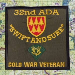 May include: A military patch with the text "32nd ADA" and "SWIFT AND SURE" in gold lettering. The patch features a red and gold crest with three trees and a green emblem with a gold eagle. The words "COLD WAR VETERAN" are at the bottom.