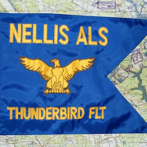 May include: A blue flag with gold embroidered text and a gold eagle emblem. The text reads "NELLIS ALS" and "THUNDERBIRD FLT". The flag is set against a map background.