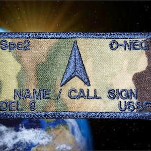 May include: Rectangular patch with a camouflage pattern, blue border, and embroidered text. The patch features a blue arrow design and the text "Spc2", "O-NEG", "NAME / CALL SIGN", "DEL 9", and "USSF".