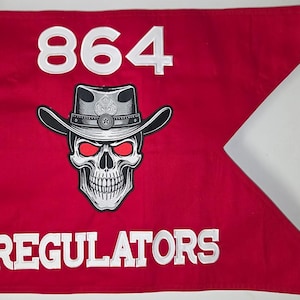 May include: Red flag with white text and a skull graphic. The text reads "864" at the top and "REGULATORS" at the bottom. The skull wears a cowboy hat and has red eyes.