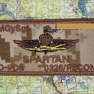 May include: A rectangular patch with the text "MGySgt" and "SPARTAN" in brown thread. The patch features a brown and gold emblem with wings and the text "0326/RECON" and "O-POS". The background has a camouflage pattern.