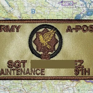 May include: Embroidered Army patch with the text "ARMY A-POS" at the top and "SGT MAINTENANCE" at the bottom. A central emblem features a brown and gold design within a black circle. The patch has a tan background and a brown border.