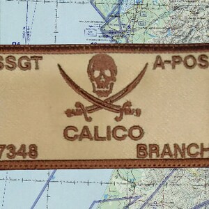 May include: A rectangular patch with a light tan background and a brown border. The patch features a skull and crossbones design above the word "CALICO". Text includes "SSGT", "A-POS", "7348", and "BRANCH".