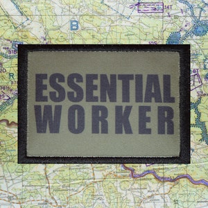May include: An olive green rectangular patch with the words "ESSENTIAL WORKER" in dark gray block letters. The patch is set against a backdrop of a detailed topographical map.