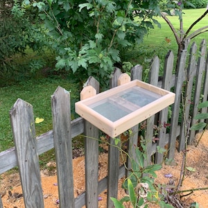 May include: A wooden bird feeder with a mesh bottom is mounted on a weathered gray picket fence. The feeder is rectangular with a light wood frame. Pink and purple flowers bloom in the background.