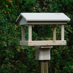 May include: A wooden bird feeder with a white roof and a platform for feeding. The feeder is mounted on a wooden pole.