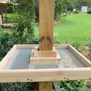May include: A wooden bird feeder with a mesh bottom to prevent seed waste. The feeder is mounted on a wooden post and has a square shape.