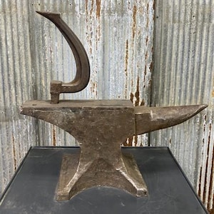May include: A weathered, antique anvil with a curved metal tool resting on top. The anvil is dark gray and shows signs of wear, with a pointed horn on one end. The tool is a darker shade of gray and has a curved handle.