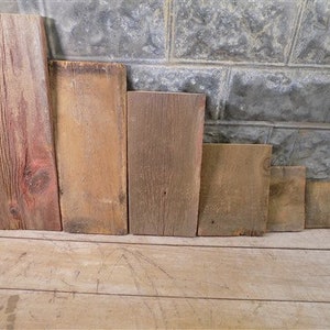 Może przedstawiać: Zestaw siedmiu odzyskanych drewnianych desek w różnych rozmiarach i odcieniach brązu, z jedną deską o czerwonawej barwie.