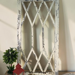 May include: A distressed, rectangular, white-painted window frame with a diamond lattice design. The frame has visible wear and tear, with peeling paint revealing the underlying wood. A red lantern and a small potted plant are in the foreground.
