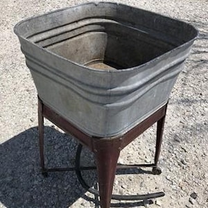 May include: A square galvanized metal wash tub with horizontal ribbed sides, supported by a dark brown metal stand featuring four angled legs and a lower cross-brace frame. The basin has an open top and a deep interior, showing a weathered finish.