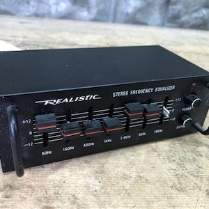 Puede incluir: Un ecualizador de frecuencia estéreo Realistic negro con controles deslizantes y perillas. La cara de la unidad tiene la palabra "Realistic" en escritura blanca, junto con marcas de frecuencia y etiquetas de control. La unidad tiene un asa a cada lado. Medidas en cm.