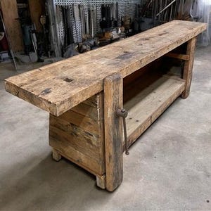 May include: A large, weathered wooden workbench with a long, rectangular top and a lower shelf. The workbench has a sturdy, aged appearance, with visible wood grain and a metal handle on the side. Ideal for woodworking projects.