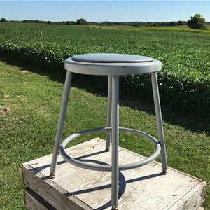 May include: A gray metal stool with a round seat and a circular footrest. The stool is sitting on a wooden surface.