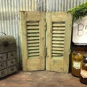 May include: A set of three vintage items: a metal box with drawers, a pair of wooden shutters, and two glass bottles. The shutters are painted green and have a distressed look. The bottles are amber glass and have labels. The metal box is painted green and has a distressed look. The items are arranged on a wooden surface.