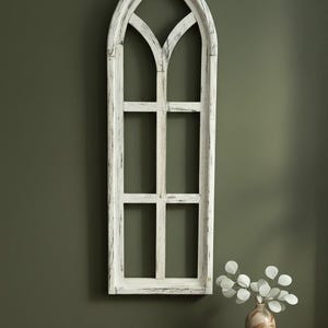 May include: A distressed white wooden wall decor in the shape of a gothic window. The window has a tall, arched top and six rectangular panes. A small vase with white decorative leaves sits on a dark wooden surface.