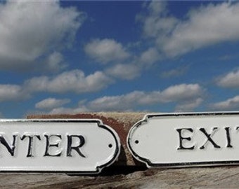May include: Two white rectangular signs with black borders and black lettering. The sign on the left says "ENTER" and the sign on the right says "EXIT".