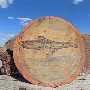 Puede incluir: Una placa redonda de madera con una imagen pintada a mano de un avión de combate P-51D en vuelo. El avión está pintado en camuflaje verde oliva y verde claro con una estrella blanca en el fuselaje. La placa está rodeada por un borde marrón y tiene el texto "P-51-D" y "8TH. U.S. AAF." inscrito en ella.