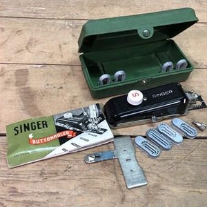 Op de afbeelding: Een vintage Singer knoopsgat naaimachine in een groene koffer met een witte knop en een rode "S" aan de bovenkant. De machine wordt geleverd met verschillende accessoires en een handleiding.