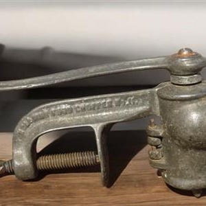 Może przedstawiać: Antyczny ręczny młynek do mięsa z żeliwa z drewnianym uchwytem. Młynek ma zacisk do mocowania do powierzchni i korbę do obracania mechanizmu mielącego.