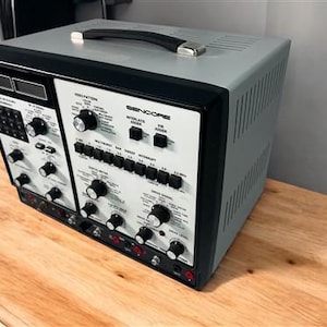 Puede incluir: Un generador de patrones de vídeo Sencore vintage con un exterior gris y negro. El dispositivo cuenta con numerosas perillas, botones y un asa. El panel frontal muestra texto como "Video Pattern" y "Sencore". Se encuentra sobre una superficie de madera.