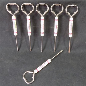 May include: A collection of six vintage bottle openers. Each opener has a silver metal loop handle and a white cylindrical body with red text. The openers are arranged on a dark surface.