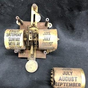 May include: An antique mechanical calendar with brass and metal components. The calendar displays days of the week and months, with visible text including "Friday," "June," and "July." The design features rotating cylinders and a circular base.