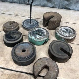 May include: A collection of antique cast iron weights in various shapes and sizes, including round, cylindrical, and a hook-shaped weight holder. The weights show signs of age and wear, with a rustic, weathered appearance.