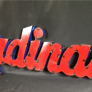 May include: Red metal sign with the word "Cardinals" in white letters with a blue outline. The sign is slightly distressed.