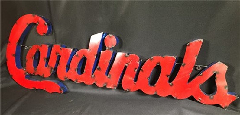 May include: Red metal sign with the word "Cardinals" in white letters with a blue outline. The sign is slightly distressed.