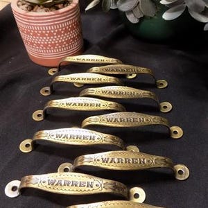 May include: Seven gold-toned metal drawer pulls with the word "WARREN" engraved on each. The pulls are arranged in a staggered pattern on a dark surface.