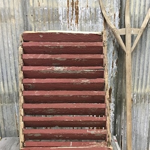 May include: A weathered red wooden shutter with multiple slats. The shutter is leaning against a corrugated metal wall.