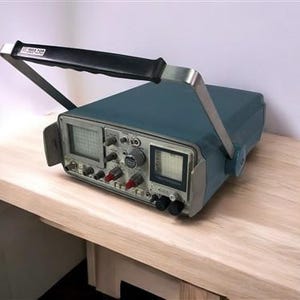 Może przedstawiać: Vintage oscyloskop w kolorze morskim z czarnym uchwytem i srebrnymi akcentami. Urządzenie posiada wiele pokręteł, przycisków i ekranów, co sugeruje jego zastosowanie w elektronice lub inżynierii. Uchwyt jest oznaczony białym tekstem. Stoi na jasnej drewnianej powierzchni.