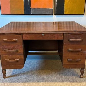 May include: A vintage wooden desk with a rich, warm brown finish. The desk features multiple drawers on each side, with curved handles. The desk has a central drawer with a keyhole and is supported by four rounded legs. The desk is a classic piece of furniture.