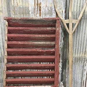 May include: A rustic wooden shelf with red paint and a weathered finish. The shelf has multiple compartments and is leaning against a corrugated metal wall.