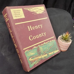 May include: A maroon book titled "Henry County" with gold lettering and a landscape illustration. The book's spine reads "Illinois" vertically. A small potted succulent plant with a pink and white patterned pot sits beside the book.