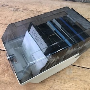 Peut inclure: Un boîtier de stockage de disquettes vintage, blanc cassé, avec un couvercle teinté transparent. Le boîtier est rempli de disquettes noires et d'un papier blanc. Le couvercle est articulé et possède une poignée. Le boîtier repose sur une surface en bois.