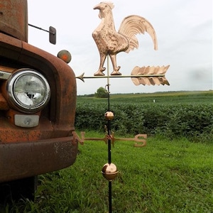 Puede incluir: Un veleta de cobre en forma de gallo con un poste de metal negro y una rosa de los vientos. El veleta está montado en un camión.