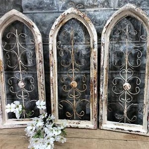May include: Three arched window frames with a distressed white and brown finish. Each frame contains a central vertical metal rod with symmetrical, swirling wire scrollwork and a small circular accent piece in the center. White flowers are placed in front.