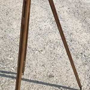 May include: A tall, vintage-style wooden tripod featuring three long, slender legs with a weathered brown finish. The legs are connected at the top by a circular metal mounting plate, creating a stable, three-legged structure for equipment support.