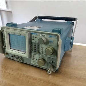 Può includere: Un oscilloscopio Tektronix 466, un dispositivo elettronico blu e grigio con uno schermo che mostra una griglia. È dotato di manopole, pulsanti e una maniglia. Il dispositivo è su una superficie di legno.