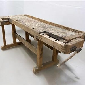 May include: A weathered wooden workbench with a rectangular top and sturdy legs. The workbench features a vise on one end and several holes along the top surface. The wood has a natural, aged appearance, suggesting it has been used for woodworking.